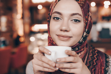 beautiful Arab girl in oriental scarf is drinking aromatic coffee in cafe and smilingの写真素材