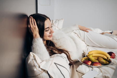 happy long-haired girl lying in bed, resting after work, next to a plate with useful fruitの写真素材
