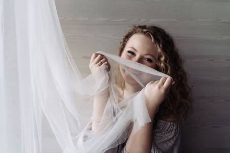 laughing young curly woman sitting by the window playing with a white curtainの写真素材