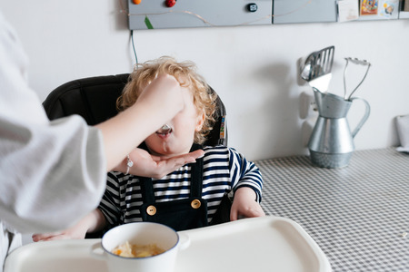 a caring young mother feeds her little curly son with useful porridge, a family breakfastの写真素材