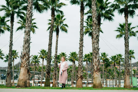 stylish girl in a trendy pink coat walks in a park next to tall green palmsの写真素材