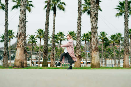 happy woman in a stylish pink coat walking in a park next to green palms, enjoying spring and warmthの写真素材