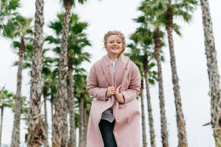 happy young woman in a trendy pink coat walking in the park, enjoying the high green palms and warmthの写真素材