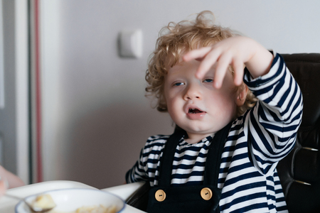 small curly baby boy sitting in a highchair in the kitchen and eating breakfast porridgeの写真素材