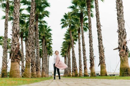 stylish young girl in a pink coat walks in the park on a warm spring day, against a background of tall green palmsの写真素材