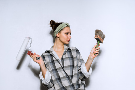 a pensive young girl in a checkered shirt holds a brush and a roller for painting the walls and can not chooseの写真素材