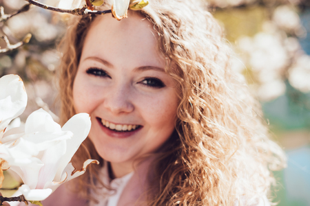 happy young woman with curly hair, in a pink coat, enjoying fragrant magnolia, in the parkの写真素材