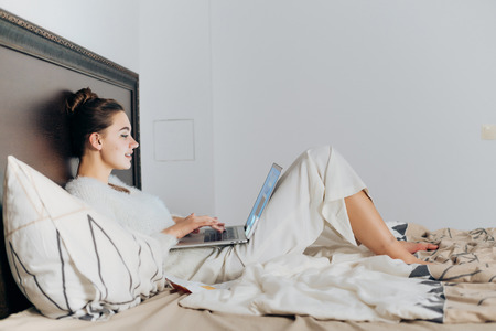 cute young girl in white pajamas lies in bed, resting and watch film on her laptopの写真素材