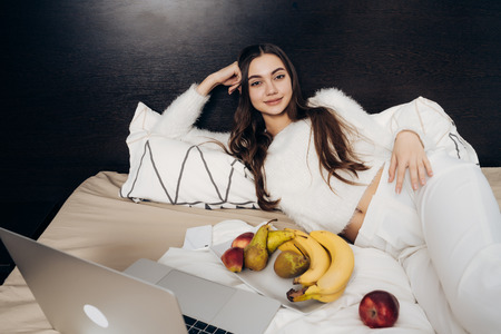 cute sleepy girl in pajamas lying in bed, resting after work, watching film on laptopの写真素材