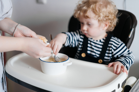 small curly baby boy sitting in a baby chair in the kitchen, family breakfastの写真素材