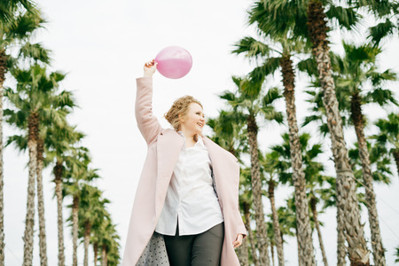 happy elegant curly woman in a coat holds a pink air balloon in her hands, walks along the green spring parkの写真素材