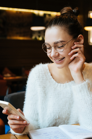 happy young girl student in glasses and a white jacket is sitting in a cafe after studying at university, looking into a smartphone and smilingの写真素材