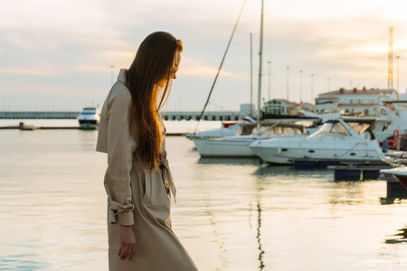 long-haired young girl in a long beige coat is standing in the seaport, waiting for her yachtの写真素材