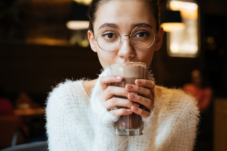 cute young girl student in glasses drinks a delicious latte in a cafeの写真素材