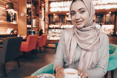 beautiful Arab girl in hijab sits in a cozy restaurant and drinks fragrant coffeeの写真素材