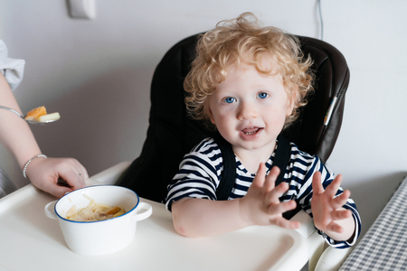 curly blonde blue-eyed boy sitting in a chair in the kitchen, waiting for a meal and smilingの写真素材