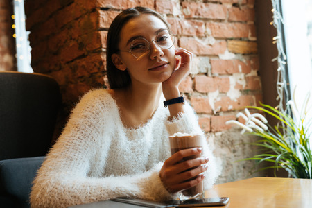 beautiful young woman in glasses sits in a cafe and drinks a delicious latte, thinks about workの写真素材