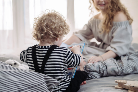 curly little baby boy sitting on the bed and playing next to mom having funの写真素材