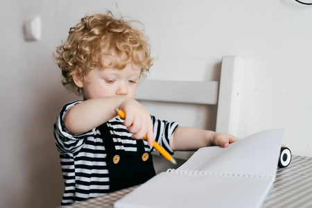 beautiful curly baby boy in cute clothes draws in a notebook, studies the worldの写真素材