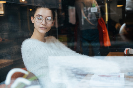 pensive young girl in a white sweater and glasses is sitting in a cafe, resting and looking out the windowの写真素材