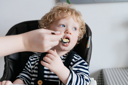 cute blonde blue-eyed boy sits in the kitchen, eats useful porridge along with his motherの写真素材
