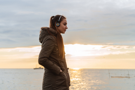 cute young girl listening to music on headphones, walking by the sea in the eveningの写真素材