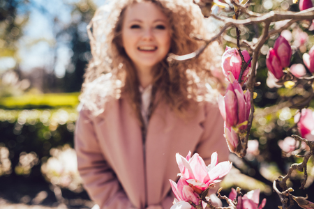curly-haired laughing woman in pink coat posing in park next to fragrant magnoliaの写真素材