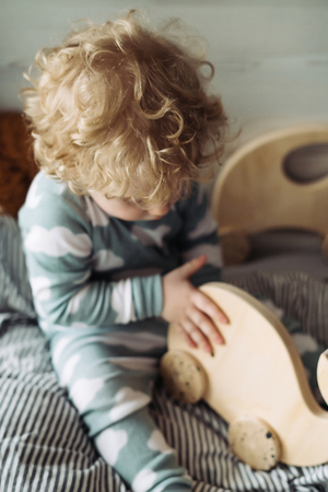 small curly blonde toddler boy in blue pajamas sitting on the bed playing with a wooden toyの写真素材