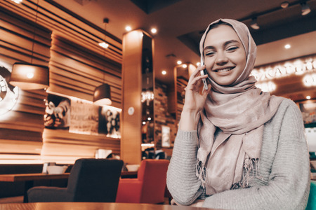 an Arab woman with a headscarf on her head talking on the phone and laughing, sitting in a cafeの写真素材