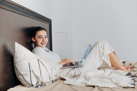 a cute smiling girl lies in bed, resting and smiling, watching a funny TV show on her laptopの写真素材