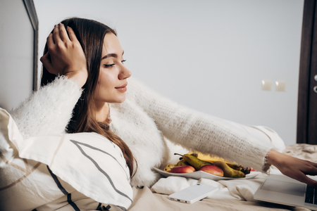 attractive young girl lies in bed at night, resting and watching film on laptopの写真素材