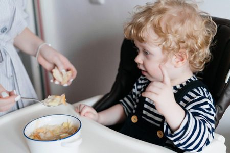 a caring young mother feeds her little curly son with useful porridge, a family breakfastの写真素材