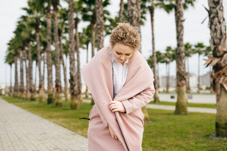 elegant young woman in a pink coat walks through the green park, enjoys the warmth and springの写真素材