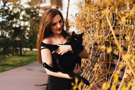 charming long-haired beautiful woman in black dress holds a black cat on her hands, walks in the garden in the eveningの写真素材