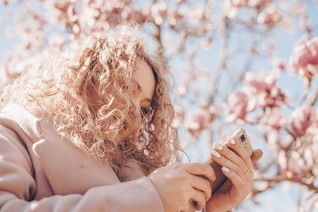 elegant curly woman enjoys fragrant magnolia in the park, looks into her smartphoneの写真素材