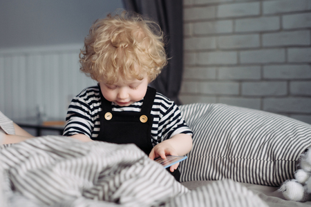 little curly baby boy in cute clothes sitting on bed and playingの写真素材