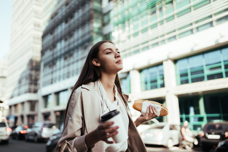 Joyful girl walks down the street and snacks on a sandwich against the background of office buildingsの写真素材