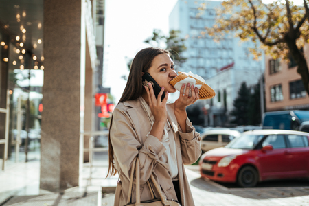 Stressed office woman eating on the way and talking on the phone outsideの写真素材