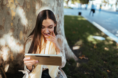 Young surprised girl looking into the tablet, sitting on the lawn, resting and watching videos online, talking in the messenger after workの写真素材