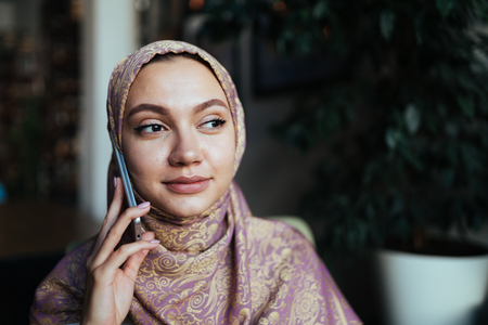 Muslim woman in a headscarf talking on the phone while sitting in a cafe, businesswomanの写真素材