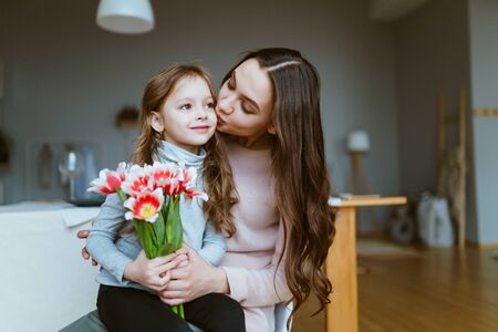 Young mother sits with her daughter on the bed and holds with her together with red tulipsの写真素材