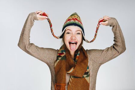 mischievously smiling girl shows tongue and holds the ends of the caps in her hands. studio gray