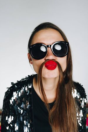 fashionable brunette with bright lipstick in sunglasses on a gray background imitates a mustache with her hair. Shockingの写真素材