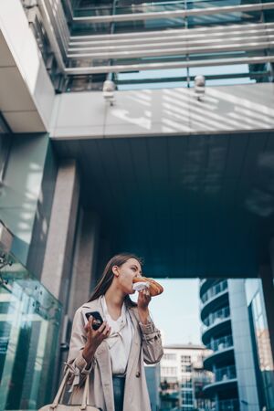 hungry-looking female student eating a hamburger and drinking coffee on the background of business buildingsの写真素材