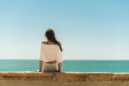 brunette sitting on the parapet with her back to the camera on the background of the ocean, legs dangling from the parapetの写真素材
