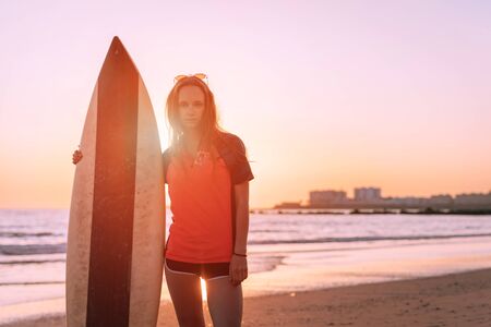 a girl with a surfboard stands by the ocean in the rays of a beautiful sunset, behind the city in...