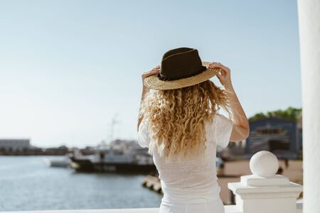 girl with curly blond hair puts on a hat on the background of ships and the sea. Girl stands with her backの写真素材