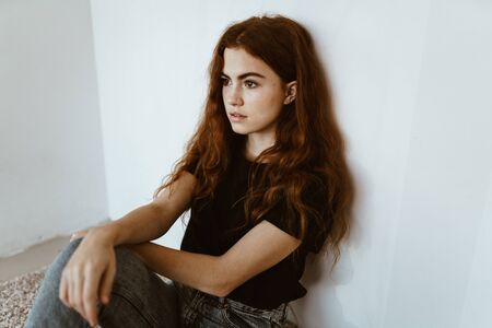 a teenage girl is sitting on the floor against the wall, her hair is scattered over her shoulders,...
