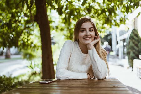 the girl smiles warmly propping her cheek with her hand and looking at the camera while sitting at the table of a street cafeの写真素材