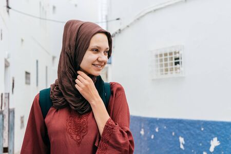 Muslim woman in a headscarf with a joyful smile holds a scarf around her neck and looks away in the...